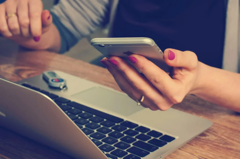 In the picture, the close-up on the hands of a woman working on her laptop. She is also holding a mobile phone in her left hand.