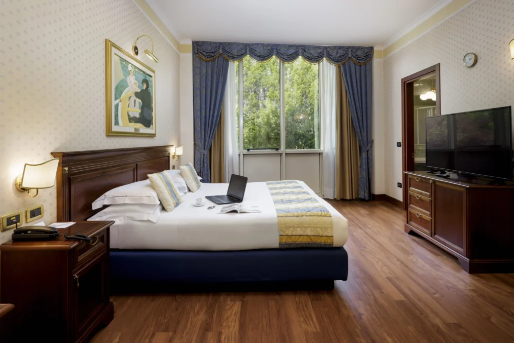 In the picture, a room of the Classic Hotel: we can see the double bed on the left and a desk with a TV on the right. ON the bed there is a laptop, a cup of coffee and an open magazine. At the back of the room, the windows from which we can see some trees outside.