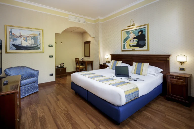 Nell'immagine, una camera del Classic Hotel: si vede il letto matrimoniale, su cui sono poggiati un laptop, una tazzina di caffè, un orologio e una rivista aperta. Di fronte al letto, si intravede un mobile con la TV e, di fianco, una poltroncina azzurra. Alle pareti sono appesi dei quadri.
