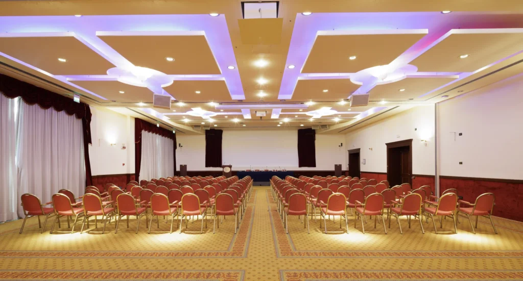 Nell'immagine, la Sala Meeting Ariosto del Classic Hotel: si vedono alcune file di sedie rosse e, di fronte, un podio con un microfono e il tavolo dei relatori. Sulla sinistra, delle ampie vetrate.